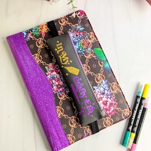 May include: A purple glitter journal with a floral and chain pattern cover, a black pencil case with the text "In My Gelly Era", and three colorful markers. The journal is open, revealing a black elastic band.