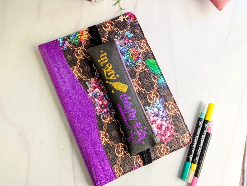 May include: A purple glitter journal with a floral and chain pattern cover, a black pencil case with the text "In My Gelly Era", and three colorful markers. The journal is open, revealing a black elastic band.