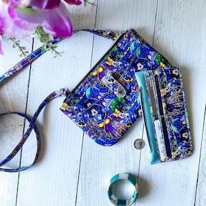 May include: A blue and purple crossbody purse with a Disney character print. The purse has a zippered pocket and a snap closure. The purse is paired with a matching blue and purple wallet with multiple card slots.