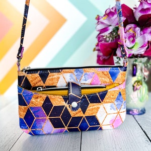 May include: A small crossbody purse with a geometric pattern in shades of blue, purple, gold, and white. The purse has a zipper closure and a flap with a snap closure. The strap is adjustable and can be worn over the shoulder or crossbody.
