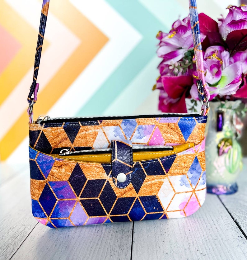 May include: A small crossbody purse with a geometric pattern in shades of blue, purple, gold, and white. The purse has a zipper closure and a flap with a snap closure. The strap is adjustable and can be worn over the shoulder or crossbody.