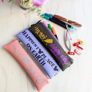 May include: A collection of pencil cases in pink, lavender, and black, each with a zipper closure and text. The pink case reads "In My Happy Place", the lavender case says "Happiest Place on Earth", and the black case says "Crafty Eie". Pens and craft supplies are scattered around.