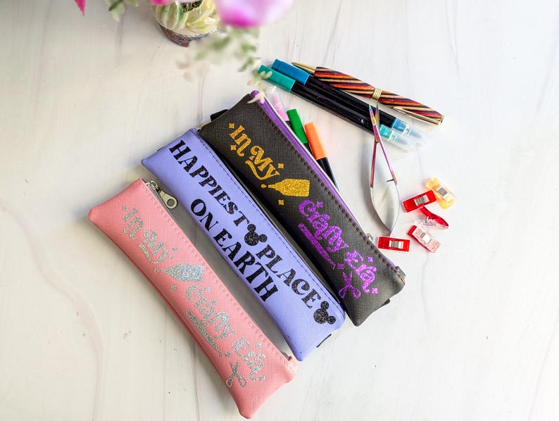 May include: A collection of pencil cases in pink, lavender, and black, each with a zipper closure and text. The pink case reads "In My Happy Place", the lavender case says "Happiest Place on Earth", and the black case says "Crafty Eie". Pens and craft supplies are scattered around.