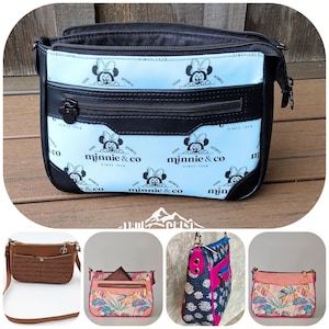 May include: A light blue crossbody bag featuring a repeating black Minnie Mouse pattern and the text "minnie & co since 1928". The bag has a black zippered front pocket with a Mickey-shaped pull, black trim, and a top zipper closure.