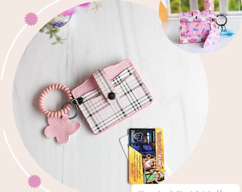 Pode incluir: Uma mini carteira rosa e branca xadrez com fecho de pressão e um amuleto de borboleta rosa preso a um anel. A carteira está sobre uma superfície branca com um cartão amarelo e branco que diz "Tickets to FUN!"