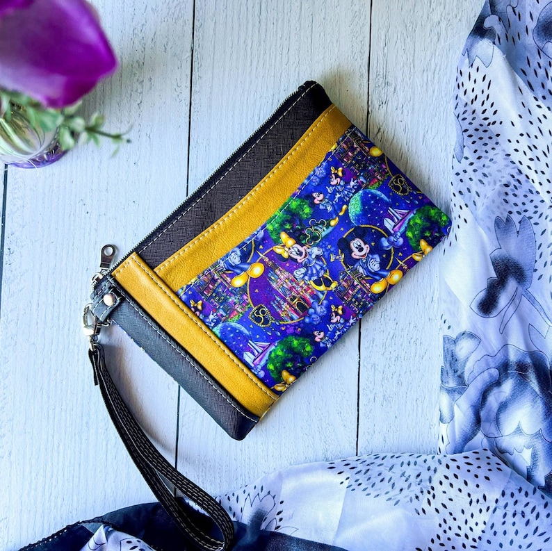 Pode incluir: Uma bolsa com z&iacute;per preta e amarela com um forro de tecido azul e roxo com uma estampa de Mickey Mouse e Minnie Mouse. A estampa inclui um castelo, fogos de artif&iacute;cio e outras imagens com tema Disney.