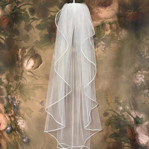 May include: A white bridal veil with a delicate, flowing design. The veil is made of sheer fabric and features a wavy, ribbon-like trim along the edges. It is displayed against a backdrop of a floral-patterned wall.