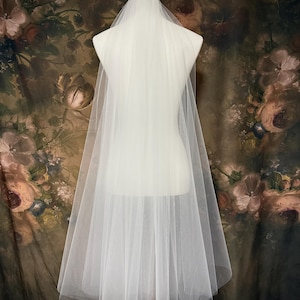May include: A sheer, floor-length white wedding veil made of fine tulle fabric. The veil is draped over a mannequin, featuring a simple, unadorned design with a soft, flowing silhouette that extends from the crown down to the floor.
