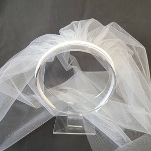May include: A clear acrylic bridal headpiece with a circular design, displayed on a clear stand. A white tulle veil is draped behind the headpiece, creating a soft, ethereal effect. The headpiece is designed for weddings and special occasions.