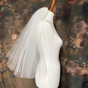 Peut inclure: Un voile de mariée en tulle blanc, transparent et multicouche, drapé sur un buste de mannequin blanc lisse et sans bras. Le voile présente une texture douce et fluide, positionné pour tomber en cascade dans le dos, révélant son tissu léger.