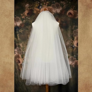 May include: A sheer white wedding veil made of fine tulle fabric. The veil is draped over a mannequin, featuring a layered design that falls in soft, vertical folds. The material is translucent, creating a delicate and airy appearance.