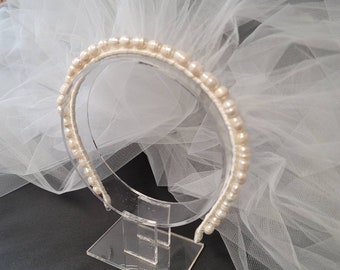 May include: A white tulle bridal veil with a pearl headband. The headband is on a clear plastic stand.