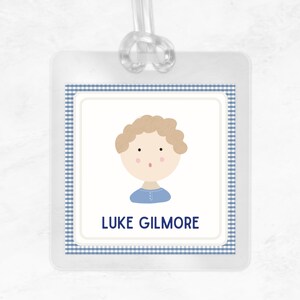 May include: A blue and white gingham patterned luggage tag with a cartoon illustration of a boy with blonde hair and a blue shirt. The text "LUKE GILMORE" is printed below the illustration.
