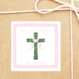 May include: A square sticker with a pink border and the words "HE IS RISEN!" above a green cross draped with a pink cloth. The text "Love, The Craigs" is below the cross. The sticker is on a brown paper package.