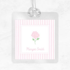 May include: A square luggage tag with a clear plastic frame and a clear loop strap. The insert features a white background with vertical light pink stripes, a central pink hydrangea illustration with green leaves, and the name Margot Smith in pink.