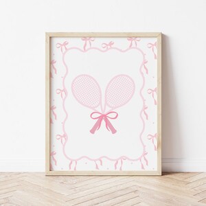May include: A framed print featuring two pink tennis rackets tied with a pink bow. The rackets are set against a white background, framed by a pink ribbon and bow border. The artwork is in a light wooden frame.
