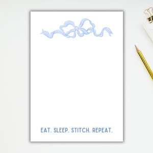 May include: A white notepad with a blue ribbon bow at the top and the text "EAT. SLEEP. STITCH. REPEAT." at the bottom.