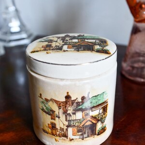 Op de afbeelding: Een cilindrische keramische pot met een afneembaar deksel, voorzien van een witte basiskleur en een gedetailleerde illustratie van een gebouw met meerdere verdiepingen, groene daken en bruine accenten op zowel de romp als het deksel, met een zilveren rand.