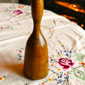 Puede incluir: Un mortero de madera vintage con una parte superior redondeada y una base acampanada. La madera tiene un tono marrón medio cálido con vetas visibles. El mortero está sobre un mantel bordado floral.