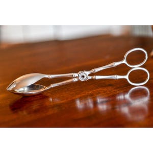 May include: Silver-colored serving tongs with a bamboo-style design. The tongs have a spoon-like scoop on one end and scissor-like handles on the other. The tongs are made of a shiny metal and are likely used for serving food.