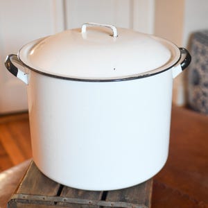 May include: A large, white enamel pot with a lid and two side handles. The pot has a black trim around the rim and handles. It sits on a wooden crate with the text "Coca-Cola Bottling Company" and the number "69" visible.