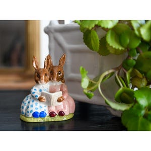 Peut inclure: Figurine en céramique représentant deux lapins bruns lisant un livre. L'un porte une robe bleue à pois blancs, l'autre une robe rose. La figurine est posée sur une base verte avec des détails rouges et bleus.