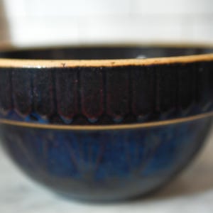 May include: A ceramic bowl with a dark blue exterior and a lighter blue interior. The rim is a darker blue with vertical ridges and a tan-colored edge. The bowl has a glossy finish.