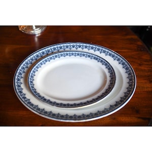 May include: Two white oval serving platters with a blue floral border are stacked on a dark wooden surface. The larger platter is beneath the smaller one. The platters are likely made of porcelain or ceramic.
