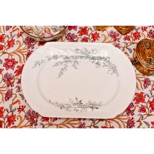 May include: An oval, off-white serving platter with a delicate floral design in gray. The platter has a slightly raised edge and is set on a floral patterned tablecloth. A small, round dish and a brass container are also visible.
