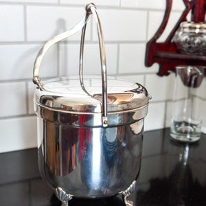 Op de afbeelding: Een glanzende zilveren metalen container met een handvat en een deksel. De container heeft een afgeronde body en staat op drie kleine, decoratieve voetjes. De handgreep buigt over de bovenkant en het oppervlak reflecteert de omgeving.