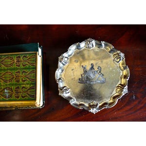 May include: A round silver-toned metal tray with a scalloped edge and a central engraved crest featuring two animals flanking a shield with the text DOMINUS AC ALVERIA. The tray has a polished finish and is positioned next to a green book.