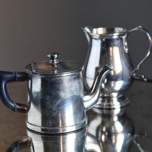 Może przedstawiać: Srebrny zestaw do herbaty z małym imbrykiem i śmietanką. Czajniczek ma czarny uchwyt i pokrywkę. Śmietanka ma zakrzywiony uchwyt. Oba elementy są odblaskowe i znajdują się na odblaskowej powierzchni.