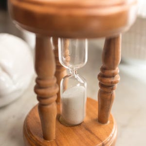 May include: A wooden hourglass with a light brown finish. The hourglass is supported by a small, round wooden stand with three curved legs. The glass chamber contains white sand.