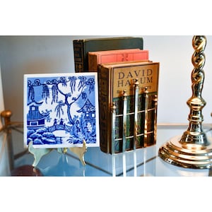 May include: A blue and white ceramic tile with a Willow pattern is displayed on a gold-coloured stand. A bookend holds several antique books. The bookend is gold-coloured with a grid design. A gold-coloured lamp base is in the background.