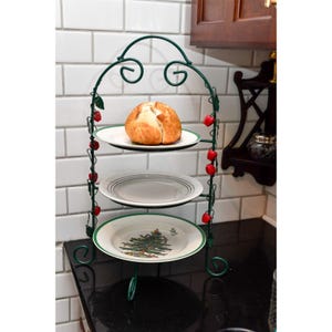May include: A three-tiered, green metal serving stand with decorative red berries and leaves. Each tier holds a white plate, with a loaf of bread on the top tier. The bottom plate features a Christmas tree design.