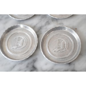 May include: Set of silver-colored metal coasters. Each coaster has a circular design with a raised edge and a large letter "D" in the center. The coasters are arranged on a marble surface.