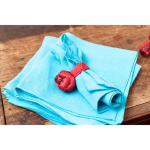 May include: A stack of light blue linen napkins with a red napkin ring. The napkin ring is a decorative knot design. The napkins are folded and arranged on a wooden surface. The image is well-lit and shows the texture of the fabric.