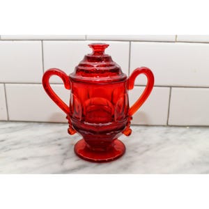 May include: A ruby red glass sugar bowl with a lid and two curved handles. The bowl has a pedestal base and is decorated with a repeating pattern. The lid has a small knob on top. The sugar bowl is sitting on a white and gray surface.
