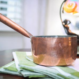 May include: A copper saucepan with a long, wooden handle rests on a green and white striped napkin. The pot has a shiny, reflective surface. The handle is a dark brown color. The saucepan is a kitchenware item.