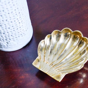 May include: A gold-toned metal dish shaped like a scallop shell with prominent vertical ridges and a smooth, reflective surface. The shell dish is positioned next to a white cylindrical object with a textured, perforated pattern.