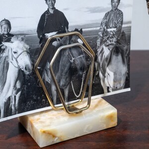 May include: A gold metal photo holder with a marble base holds a black and white photo of three people riding horses. A small brass statue of the Statue of Liberty is to the right of the photo holder.