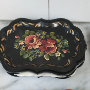 May include: Black decorative tray with a scalloped edge, adorned with a floral design featuring red and pink roses, green leaves, and small white and blue flowers. The tray has gold accents.