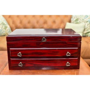 May include: A rectangular, wooden jewelry box with a glossy, reddish-brown finish. The box has two drawers with ornate, gold-colored handles and a hinged top with a clasp. The wood grain is visible, adding to the box's classic aesthetic.
