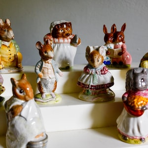 May include: A collection of ceramic figurines depicting various animals in anthropomorphic poses. The figurines include a rabbit, mouse, squirrel, frog, and pig, each dressed in detailed clothing. The figurines are painted in bright colors and are displayed on a white shelf.