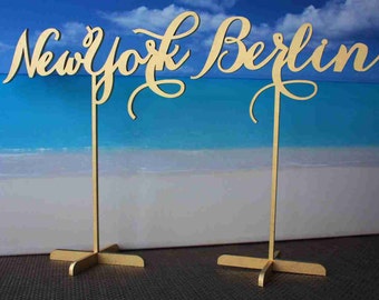 May include: Two wooden signs with the words "New York" and "Berlin" written in a cursive font. The signs are mounted on gold metal stands with a cross-shaped base.