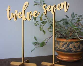 May include: Two gold wooden table number signs with the words "twelve" and "seven" written in a cursive font. The signs are on a brown surface with a potted plant in the background.