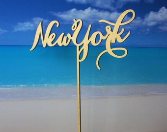May include: Gold wooden tabletop sign with the word "New York" in a cursive font. The sign is on a stand with a cross-shaped base.