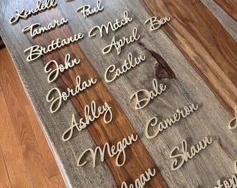 Puede incluir: Recortes de nombres dorados en fuente cursiva dispuestos sobre una superficie de madera. Los nombres incluyen Kendell, Daniel, Tamara, Paul, Brittance, Mitch, John, April, Jordan, Caitlin, Ashley, Dale, Megan, Cameron y Shaun.