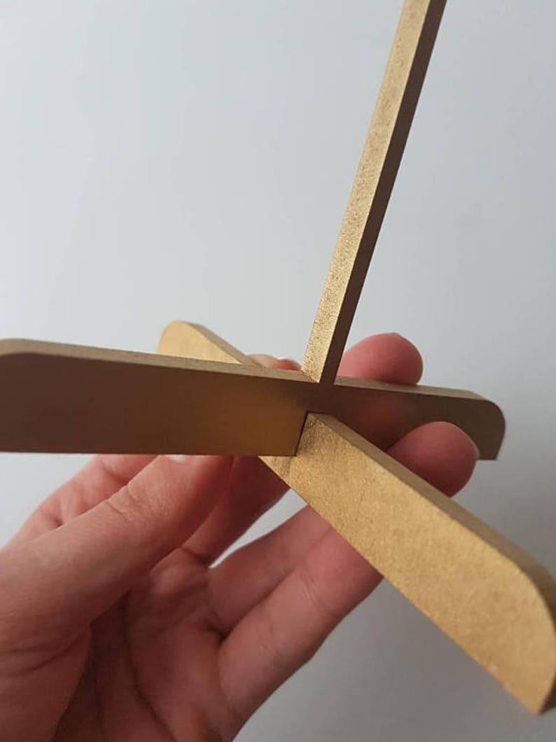 Op de afbeelding: Een gouden houten standaard met vier poten die een X vormen. De standaard is gemaakt van dunne, platte stukken hout.