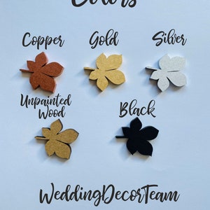 Op de afbeelding: Een set van zes houten bladeren in verschillende kleuren, waaronder koper, goud, zilver, zwart en onbehandeld hout. De bladeren zijn in een cirkel op een lichtblauwe achtergrond gerangschikt. De tekst "Colors" staat bovenaan de afbeelding, en de tekst "Wedding Decor Team" staat onderaan.